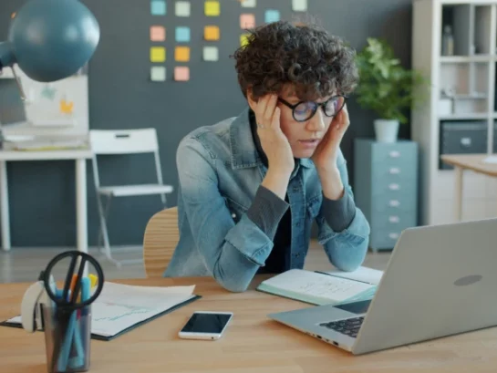A clearly tired and overworked woman sits at a computer, massaging her temples.